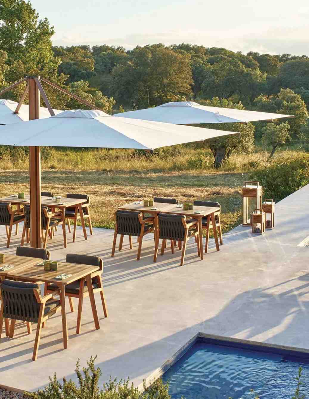 Outdoor dining area with tables, chairs, and umbrellas near a pool with a scenic background.