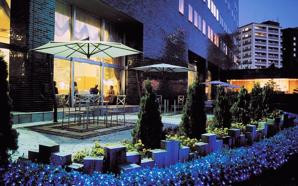 Evening outdoor setting with people sitting at tables under umbrellas, surrounded by greenery and buildings.