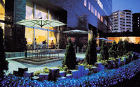 Evening outdoor setting with people sitting at tables under umbrellas, surrounded by greenery and buildings.