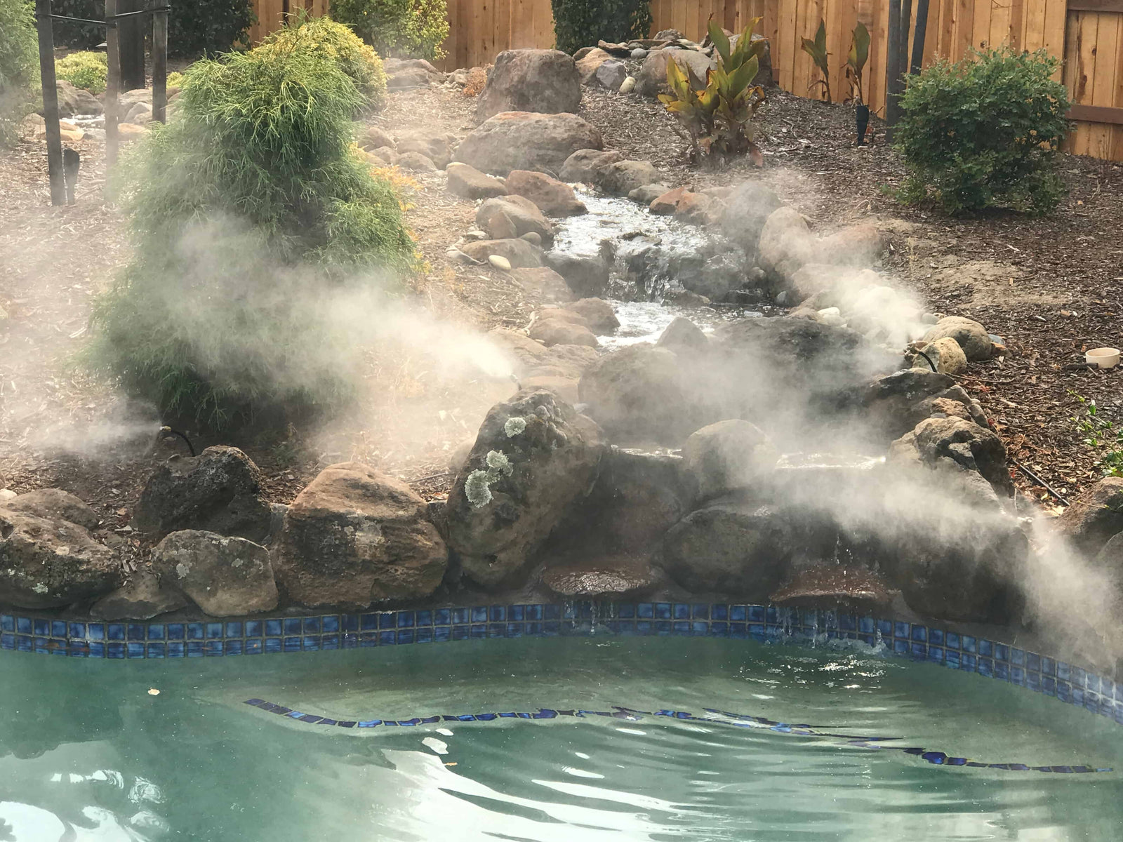 Outdoor pool with steam rising from hot tub area, surrounded by rocks and plants.