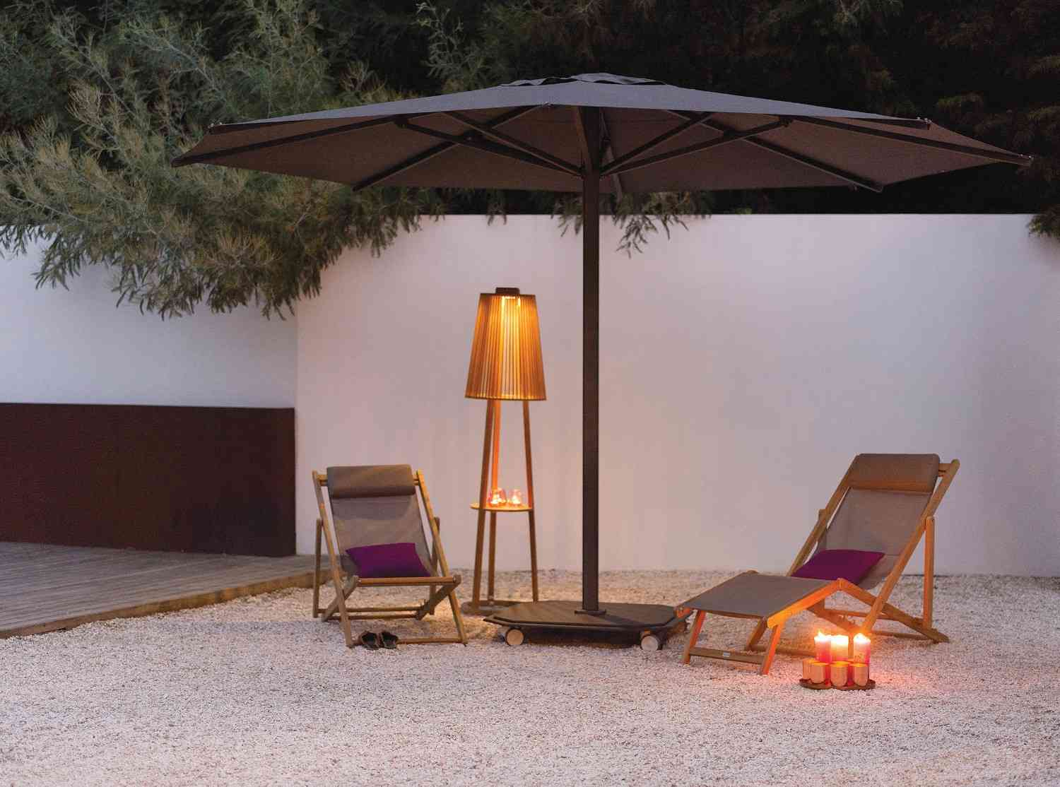Outdoor setting with two lounge chairs under a large umbrella, illuminated by a floor lamp and candles.