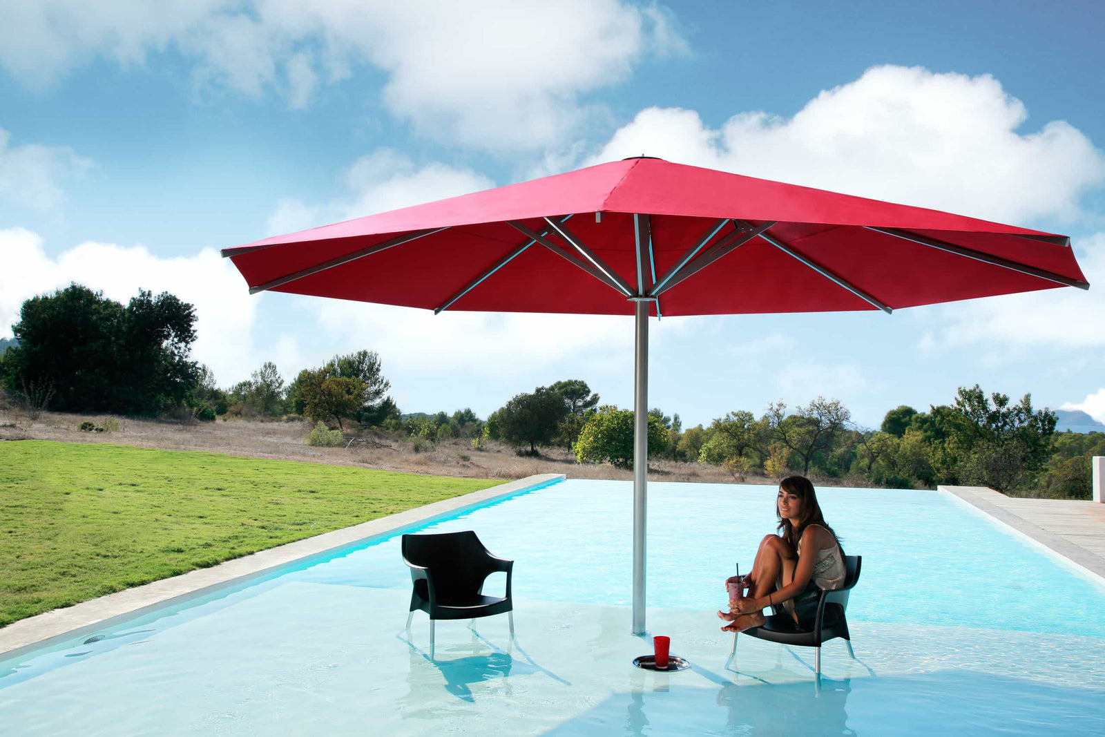 Person sitting under a large red umbrella by a pool with a scenic background