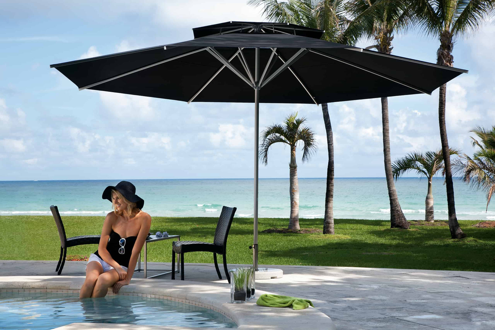 Woman sitting by a pool with a large black patio umbrella, palm trees, and ocean view.