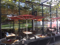 Outdoor seating area with tables, chairs, and red umbrellas in a park-like setting.