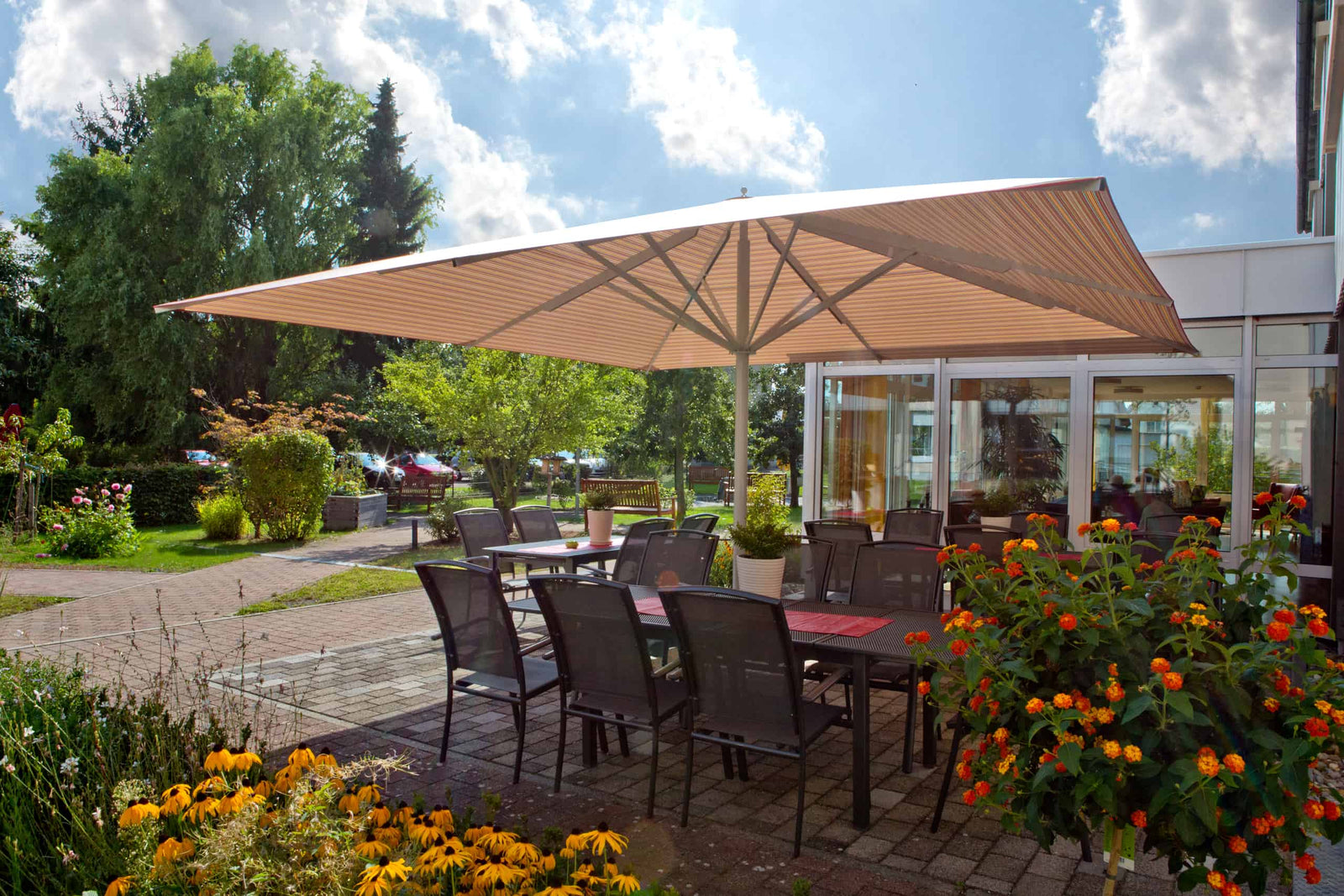 Outdoor dining area with large umbrella, tables, chairs, and flowers on a sunny day.