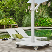 White patio umbrella with stand on a wooden deck by a poolside with lounge chairs and greenery.
